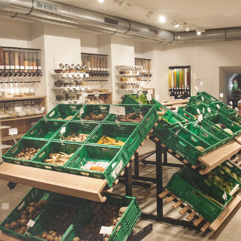 Ein Foto des Ladeninneren. In der Mitte grüne Kisten mit Obst und Gemüse. Im Hintergrund Lebensmittel-Spender zum Gefäße selbst-befüllen und die Kühlung.