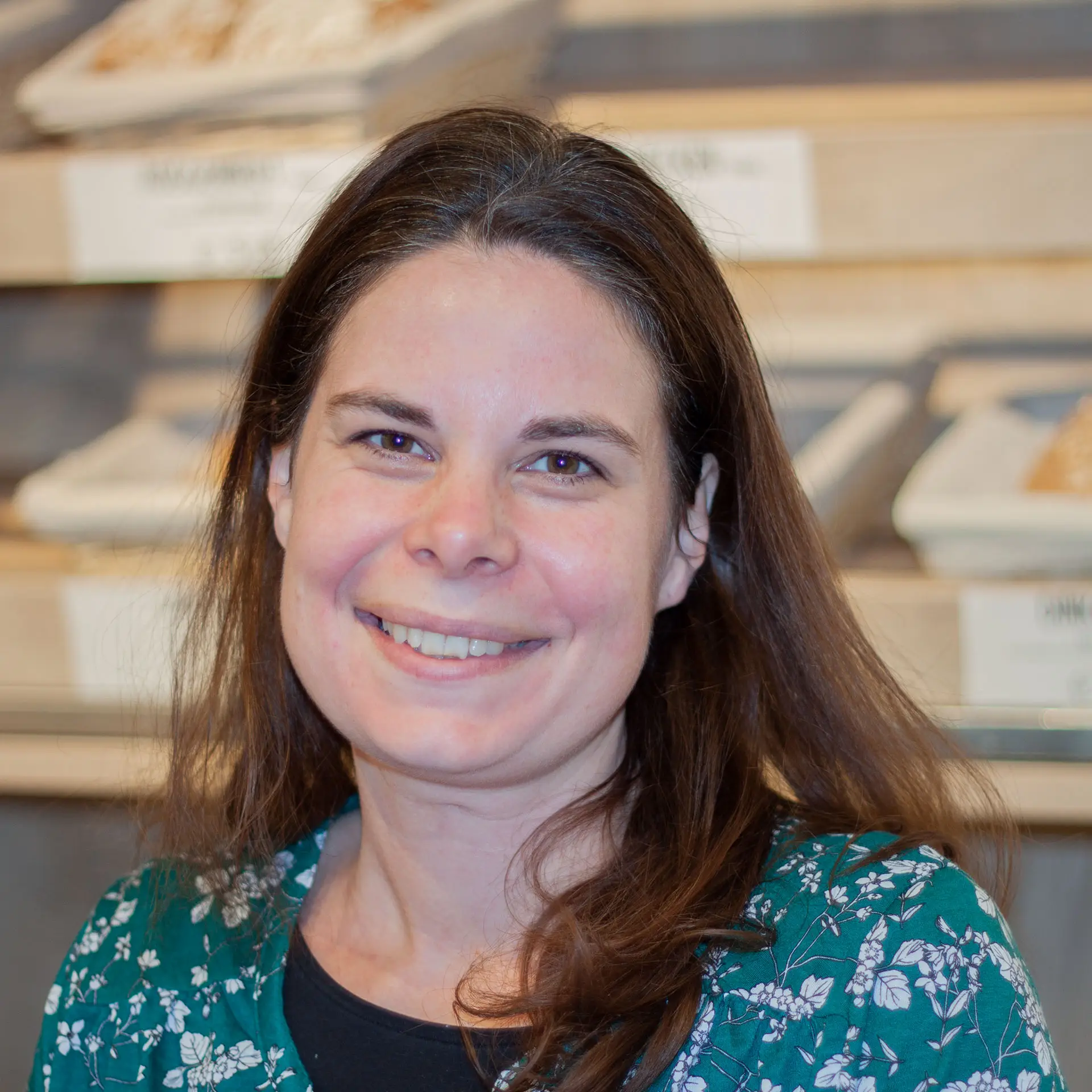 Ein Foto von Claudia. Sie hat lange braune Haare, trägt ein blau-grünes Shirt mit weißen Blumen und lächelt.